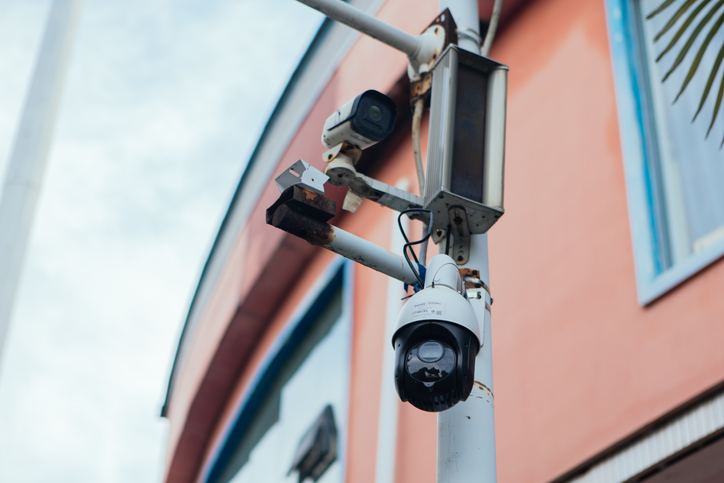 Low-angle shot of dome speed dome camera and bullet surveillance camera mounted on rusted white utility pole beside warm orange building facade.
