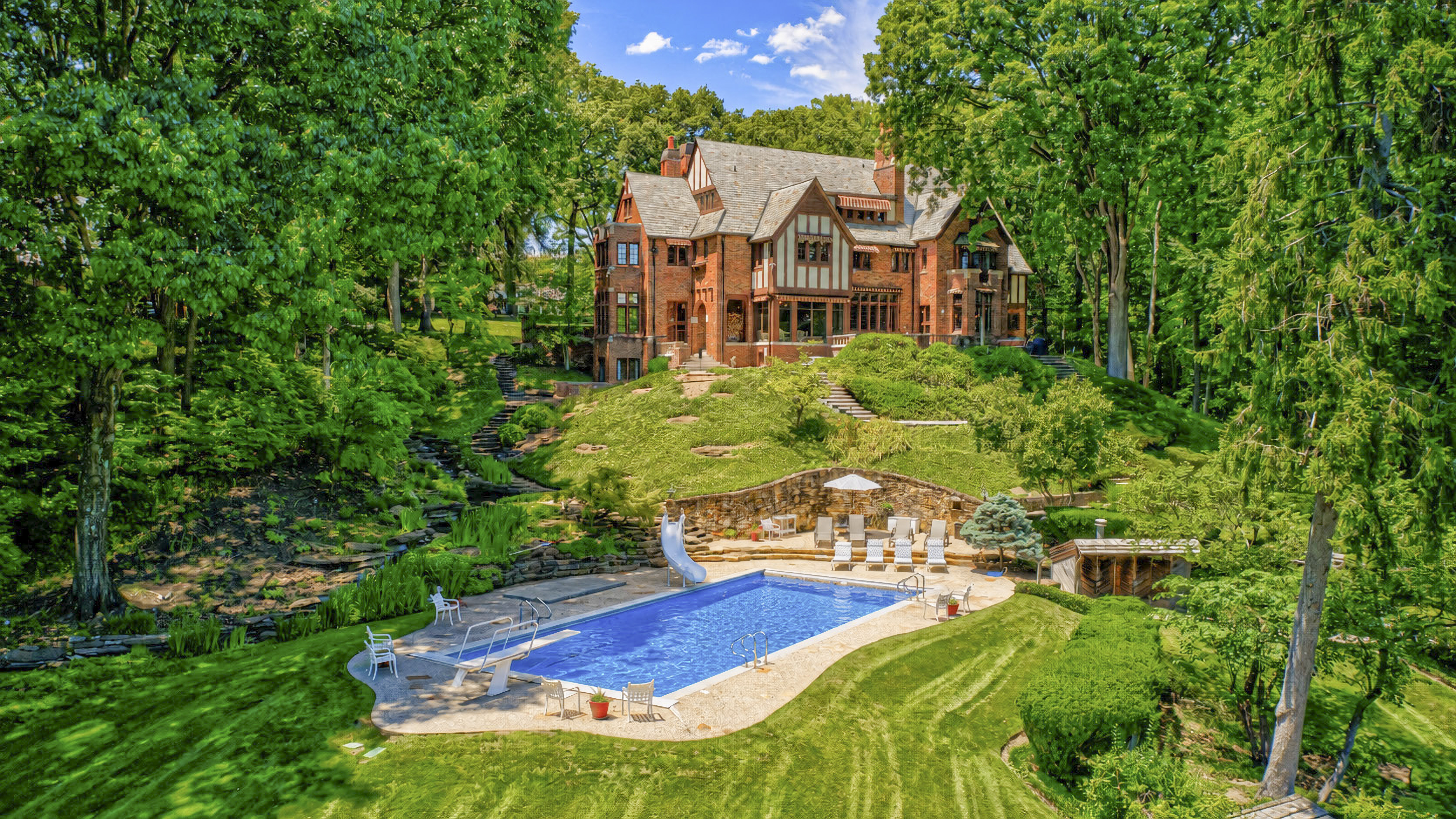 A large, ornate Tudor-style mansion surrounded by lush, verdant gardens and a swimming pool in the foreground.