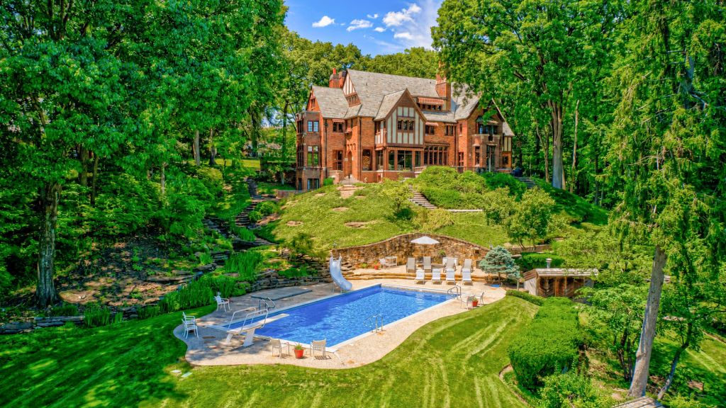 A large, ornate Tudor-style mansion surrounded by lush, verdant gardens and a swimming pool in the foreground.