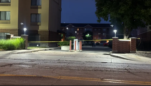 Nighttime view of a parking lot with brick buildings, trees, and lampposts illuminating the scene.