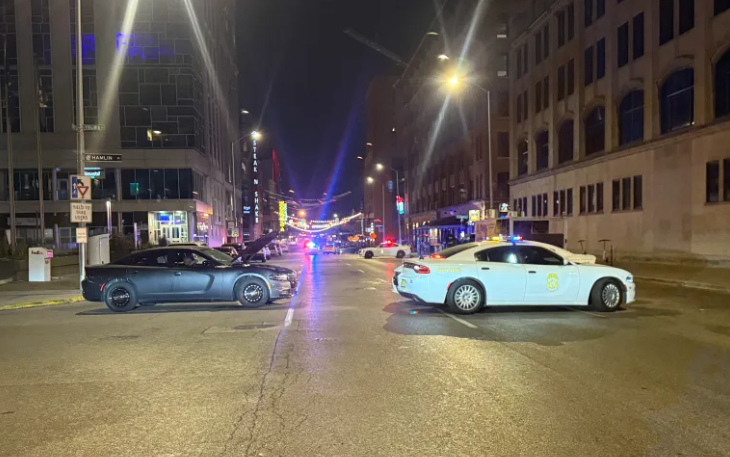 A nighttime street scene with several cars, including a taxi, parked on the road. Bright lights and buildings are visible in the background.