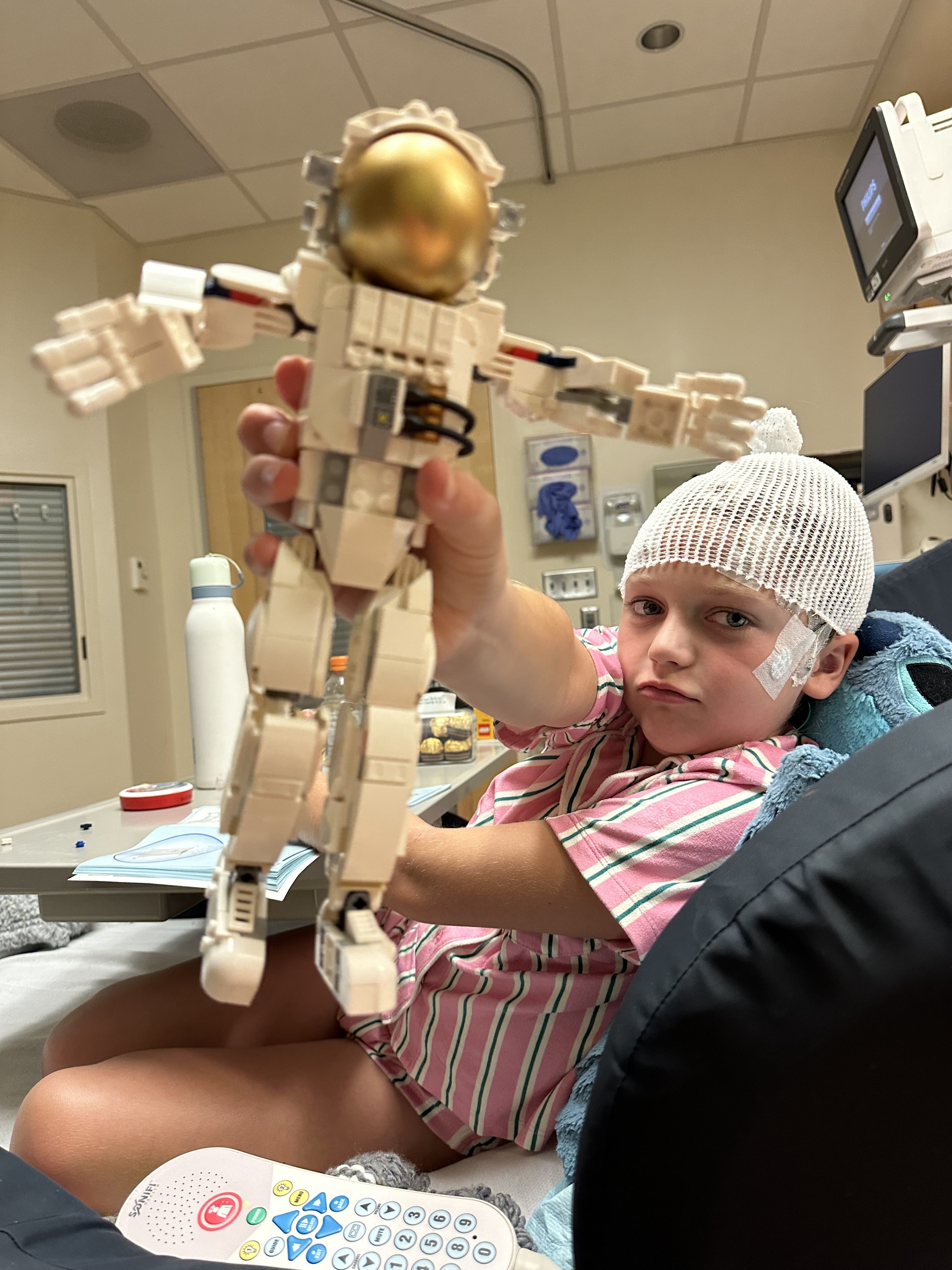 A young child wearing a pink and white striped shirt and a white hat sits in a hospital bed, holding a toy robot with a golden head.