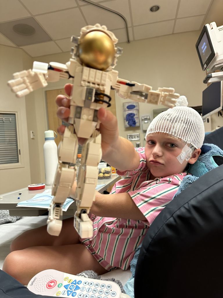 A young child wearing a pink and white striped shirt and a white hat sits in a hospital bed, holding a toy robot with a golden head.