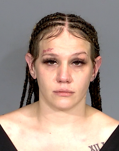 A close-up portrait of a young woman with braided hair and a serious expression, wearing a black top.
