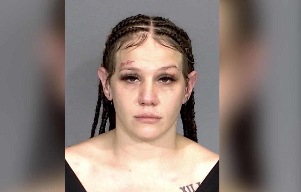 A close-up portrait of a woman with braided hair and a serious expression, wearing a black top.