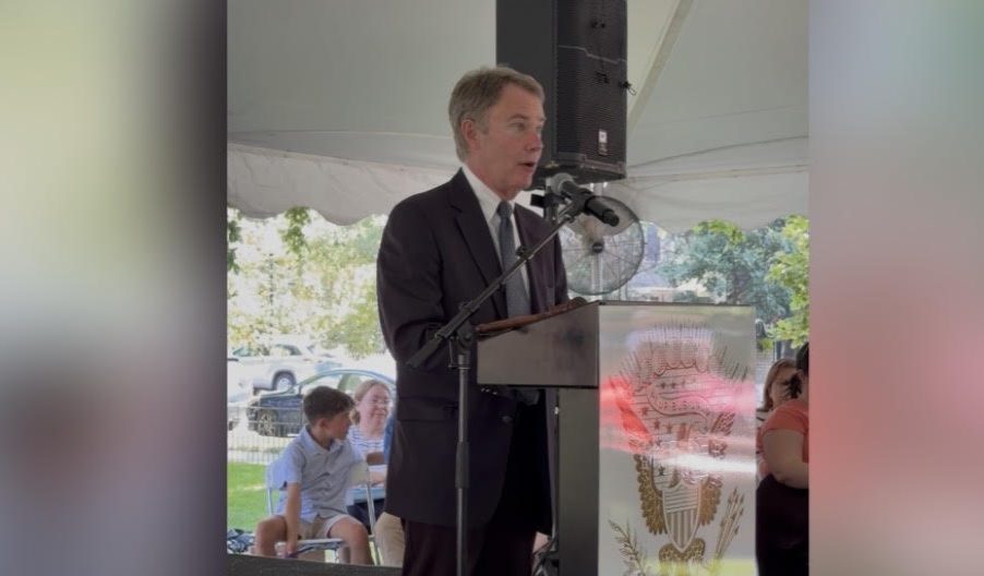 A man in a suit standing at a podium and speaking into a microphone at an outdoor event, with a banner behind him displaying the name "Benjamin Harrison".