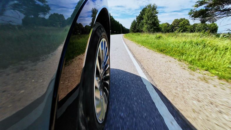 Car tire rolling along rural road with green vegetation, blue sky, and visible edge of pavement