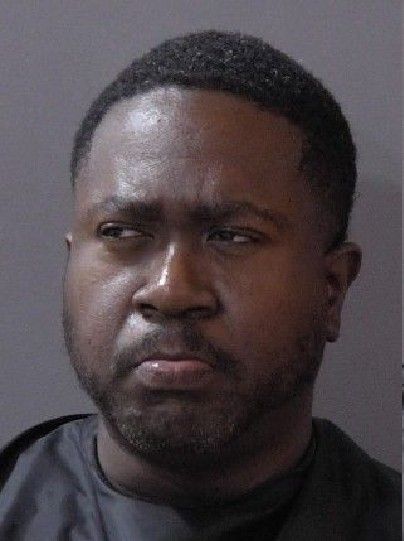 Headshot of a serious-looking Black man with a beard wearing a dark shirt against a gray background.