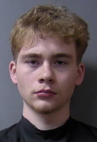 A young man with short, wavy blond hair and a serious expression, wearing a black shirt.