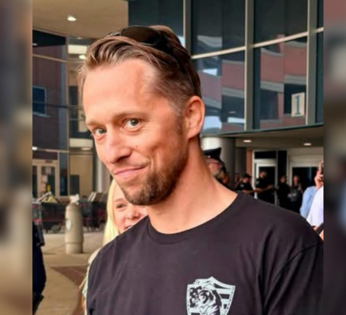 A man with a beard and long hair wearing a black t-shirt with a tiger logo.