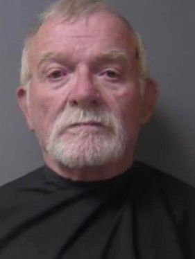 Headshot of an elderly man with a long white beard wearing a black shirt against a gray background.