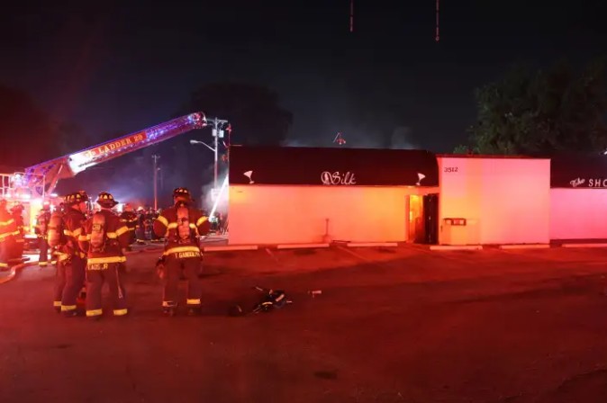 Nighttime scene of firefighters and emergency vehicles at a building fire, with "911" and "SHOP" visible on the structure.