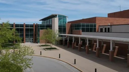 A large brick building with glass walls and entrances, surrounded by a paved plaza with benches.