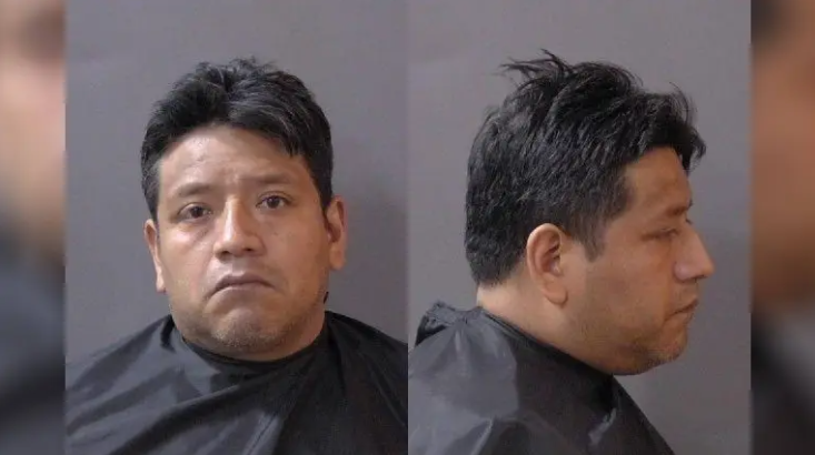 A headshot of a man with short dark hair and a serious expression, wearing a black shirt against a gray background.