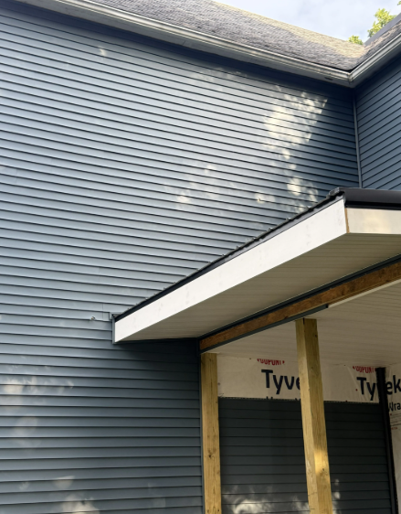 A gray siding exterior of a building with a wooden overhang and Tyvek house wrap visible.