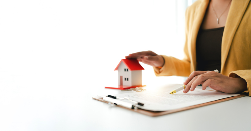 Woman's hand hovering over a small house model and contract document, representing property investment, real estate business, home buying, and financial protection concepts