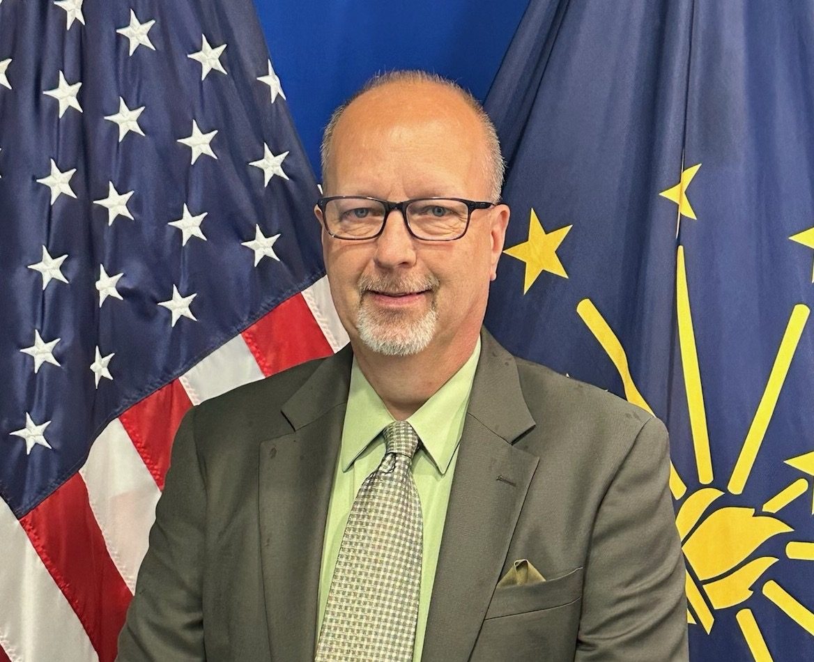 A middle-aged man with glasses and a beard wearing a suit and tie, standing in front of American and Indiana state flags.