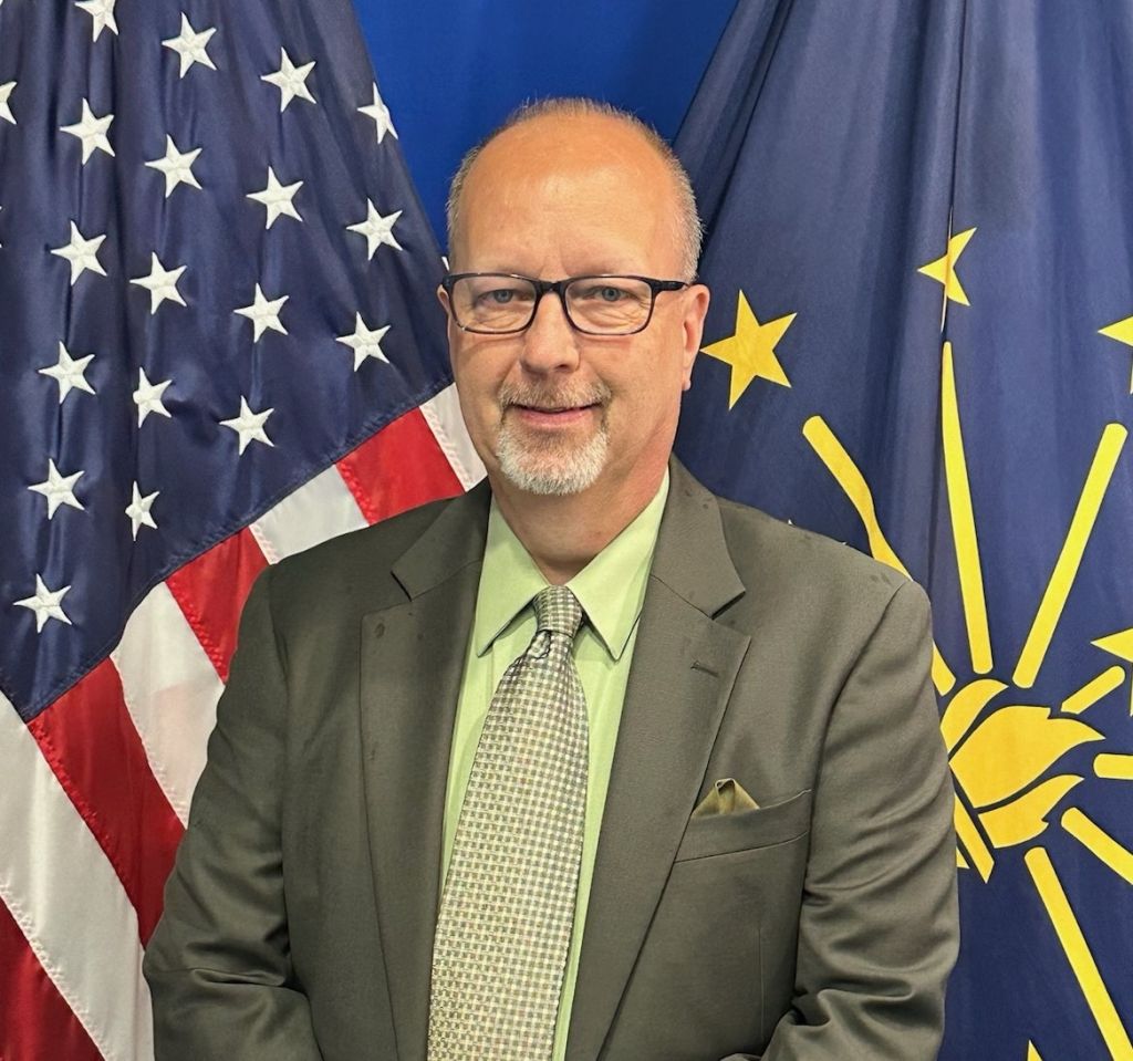 A middle-aged man with glasses and a beard wearing a suit and tie, standing in front of American and Indiana state flags.