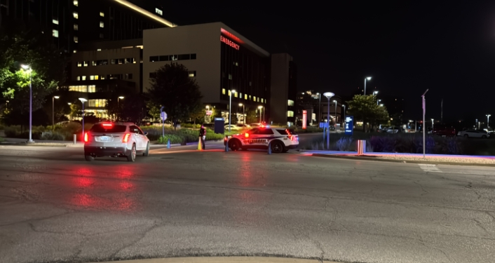 Night scene of a city street with several vehicles, including a police car with flashing red lights, parked along the curb.