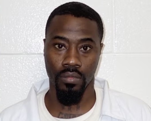 A close-up portrait of a Black man with a beard, wearing a white shirt and looking directly at the camera.