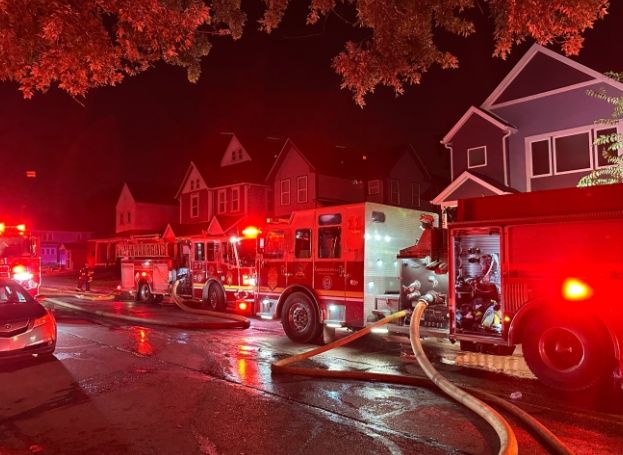Fire trucks and firefighters responding to an emergency at night on a residential street with illuminated houses and trees.