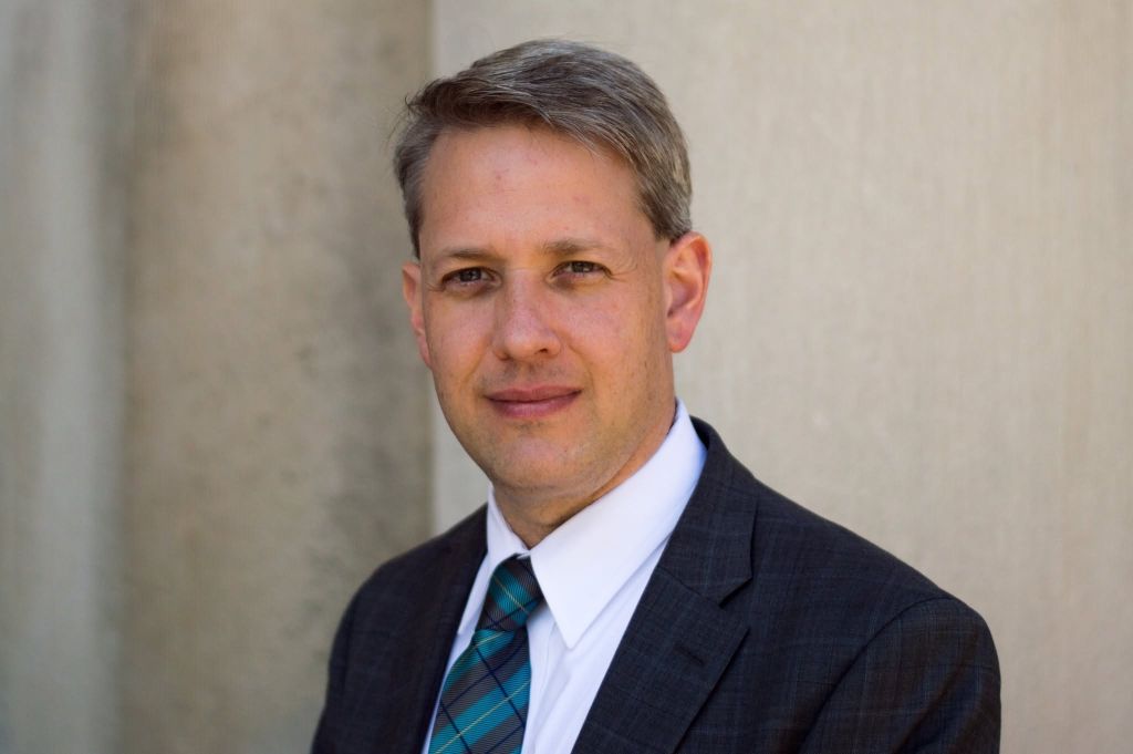 A middle-aged man wearing a suit and tie, standing in front of a plain wall and looking directly at the camera with a neutral expression.