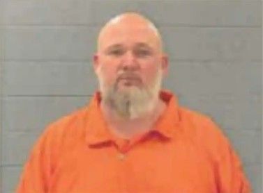 A middle-aged man with a beard wearing an orange prison uniform, standing in front of a gray wall.