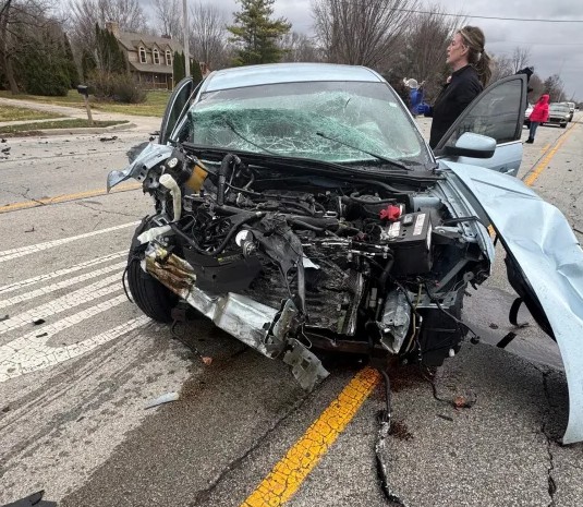 A damaged black vehicle with a shattered windshield and front end damage on the side of a road.