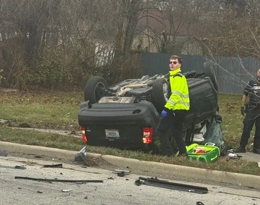 Overturned vehicle on the side of the road, with a person in a high-visibility vest standing next to it.