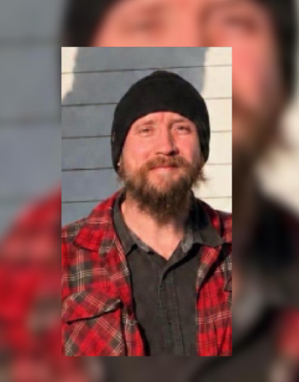 A bearded man wearing a red and black plaid shirt and a black knit hat, standing in front of a wooden wall.
