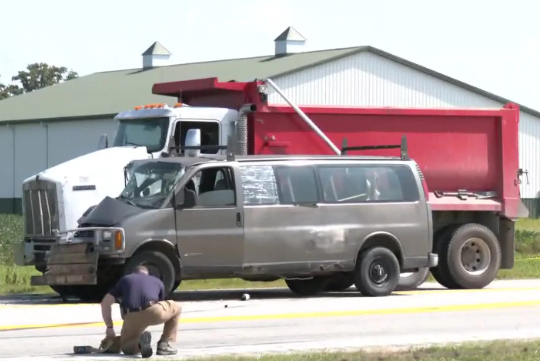 A large red dump truck parked next to a person crouched on the ground in a grassy field, with a metal building in the background.
