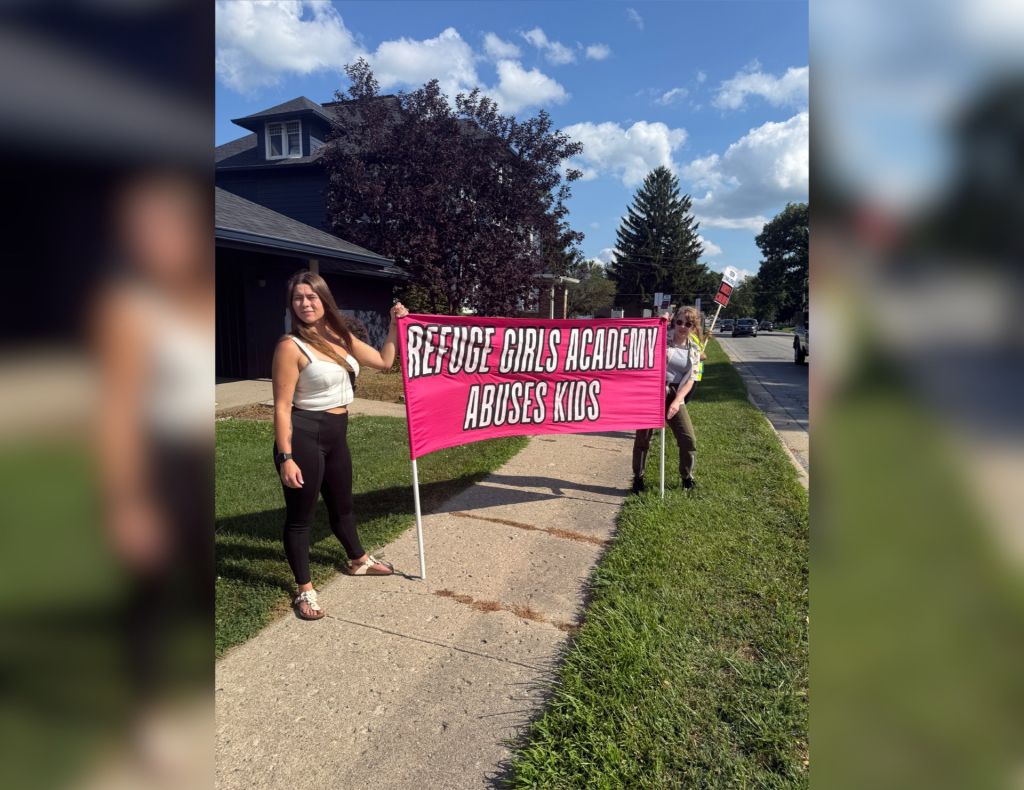 A young woman holding a large pink banner that reads "REFUGE GIRLS ACADEMY ABUSES KIDS" while standing on a sidewalk in front of a building.