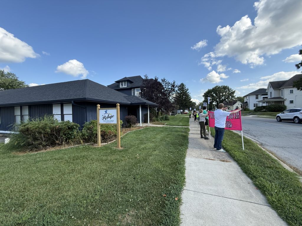 A residential neighborhood with a building labeled "Refuge" and people holding signs on the sidewalk.