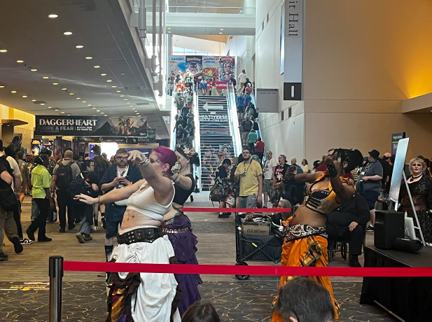 Crowded convention center with people in cosplay costumes, some dancing on stage, and Daggerheart video game banner visible.