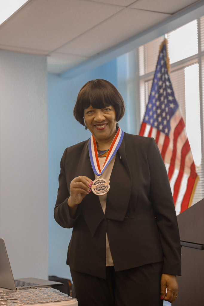 A smiling woman wearing a suit and medal stands in front of an American flag.