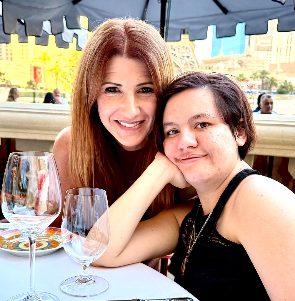 Two smiling women sitting at a table with wine glasses, in front of a city skyline backdrop.
