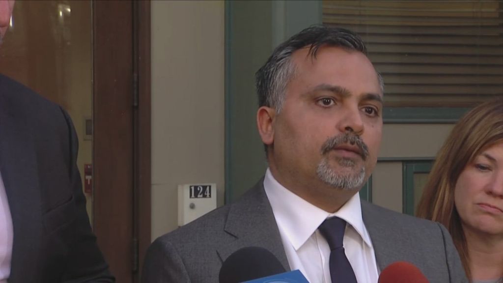 A man in a suit standing in front of a building, speaking to reporters and being filmed.