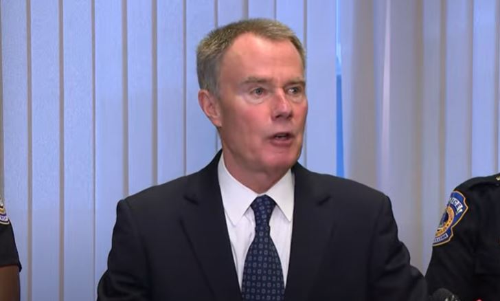 A man in a suit speaking at a podium with a police badge visible.