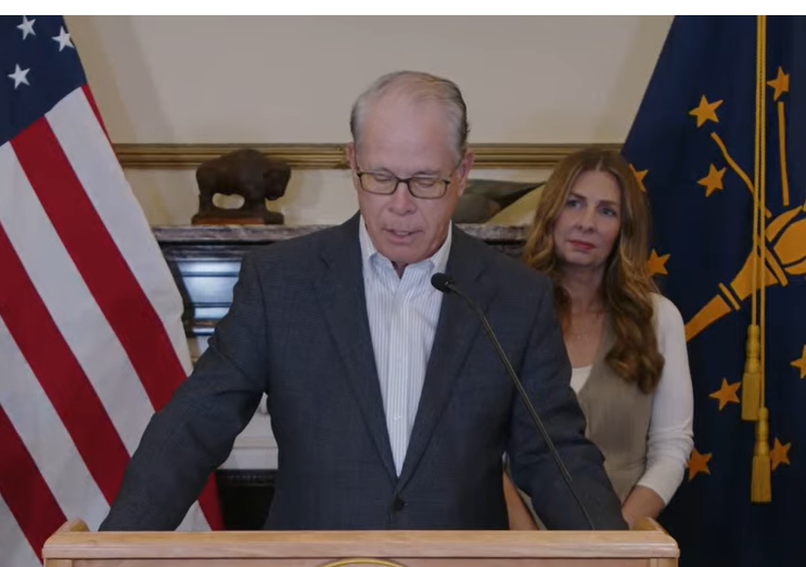 Two people, a man and a woman, standing at a podium with an American flag and state flag in the background.