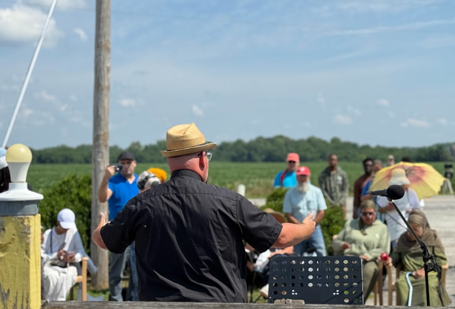 A group of people gathered outdoors, with a man in a hat speaking into a microphone on a podium.