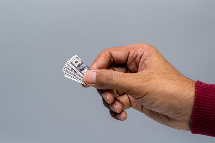Close-up of a human hand holding tiny, miniature US dollar bills against a gray background.