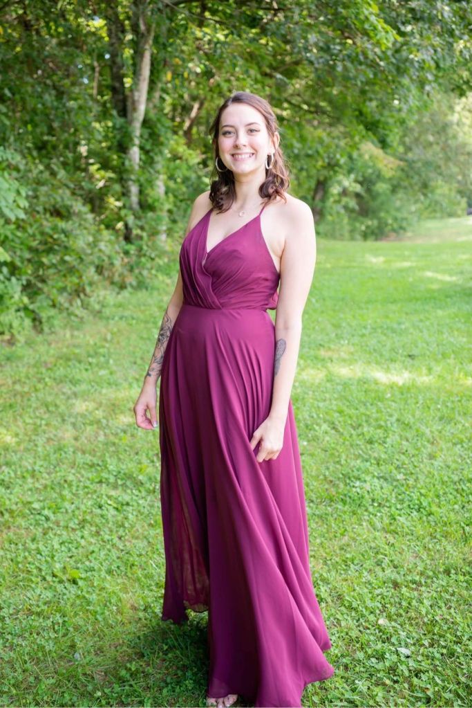 A smiling young woman in a purple dress standing in a grassy, wooded area.