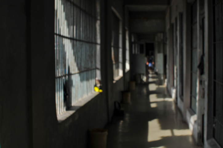 A perspective shot of a gloomy, empty prison hallway or corridor lined with heavy iron bars on the windows and cell gates, showing natural light casting shadows on the floor.