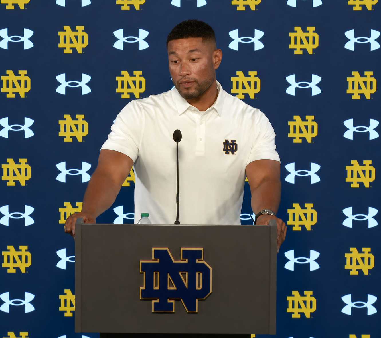 A man in a white polo shirt stands at a podium with the University of Notre Dame logo, speaking into a microphone in front of a backdrop featuring the Notre Dame logo and Under Armour branding.
