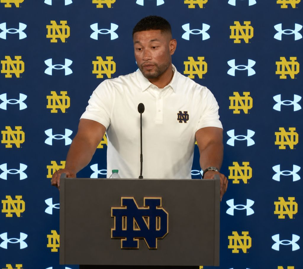 A man in a white polo shirt stands at a podium with the University of Notre Dame logo, speaking into a microphone in front of a backdrop featuring the Notre Dame logo and Under Armour branding.