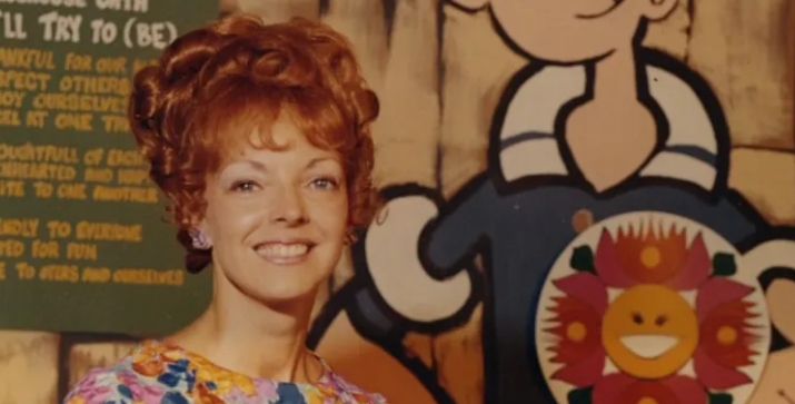 A smiling woman with curly red hair stands in front of a colorful cartoon background.