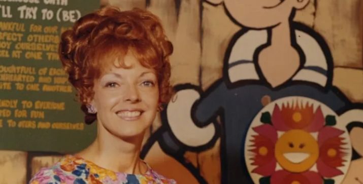 A smiling woman with curly red hair stands in front of a colorful cartoon background.