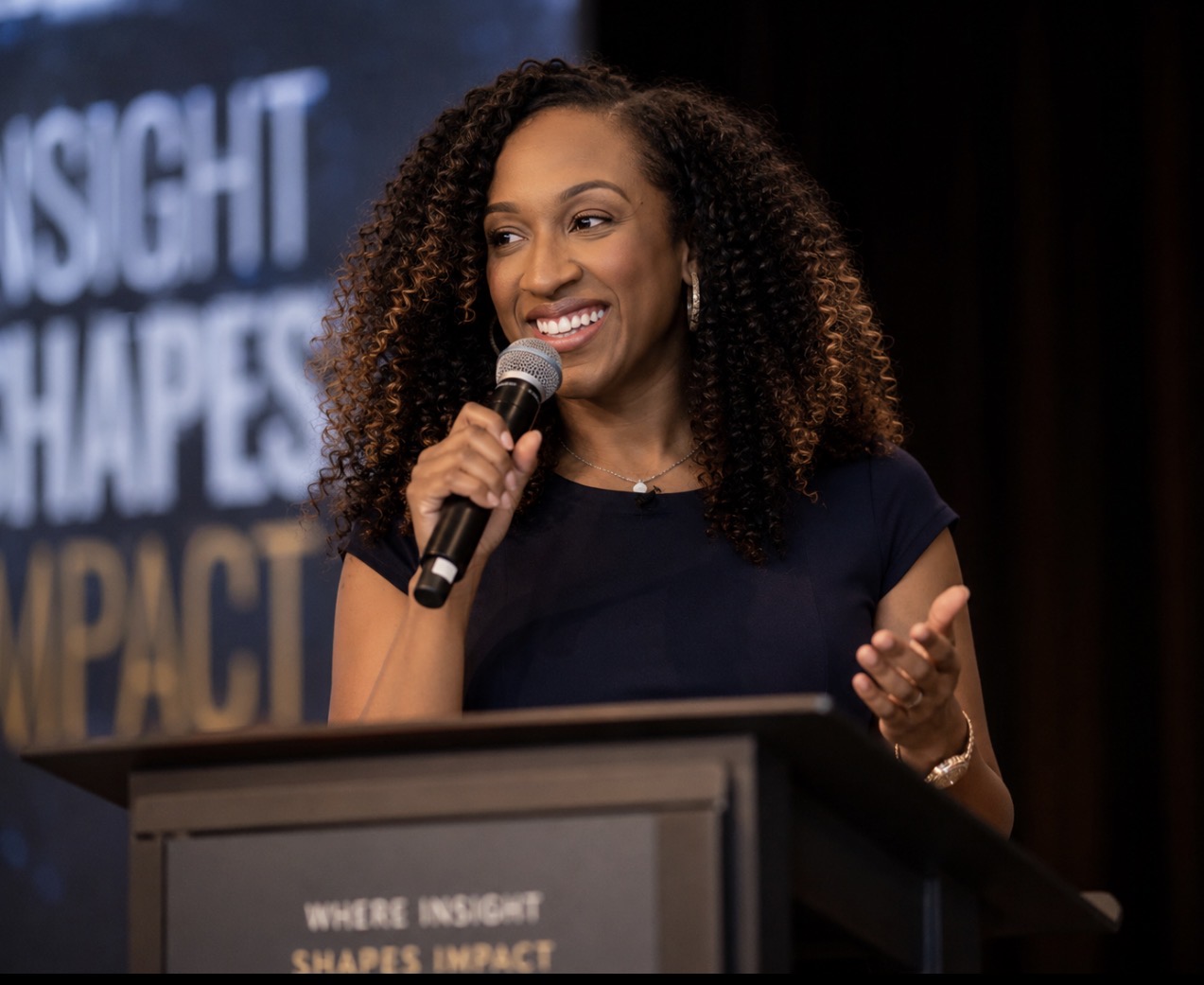 A smiling woman with curly hair speaking into a microphone at a podium with the text "Insight Shapes Impact" visible.