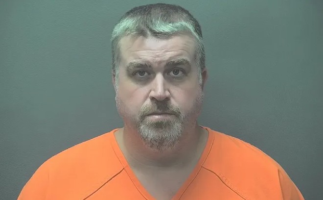 A middle-aged man with gray hair and a beard wearing an orange prison uniform, looking directly at the camera.