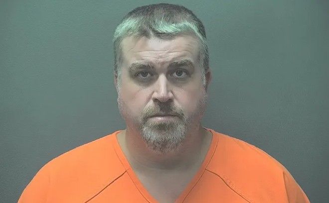 A middle-aged man with gray hair and a beard wearing an orange prison uniform, looking directly at the camera.